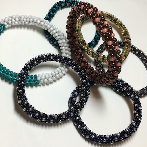 May include: A set of six beaded bracelets in various colors, including teal, white, black, brown, and green. The bracelets are made with small beads and have a braided design. The bracelets are arranged in a pile on a white surface.