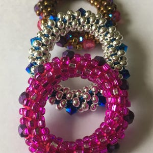 May include: Three beaded rings stacked on top of each other. The bottom ring is pink with purple accents, the middle ring is silver with blue accents, and the top ring is brown with pink accents.