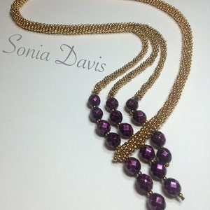 May include: A gold beaded necklace with a long, cascading strand of dark purple beads. The necklace is layered and has a delicate, feminine look.