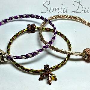 May include: Three braided bracelets in shades of purple, silver, gold, green, and brown. One bracelet has a decorative silver bead, another has a brown wooden bead, and the third has a rose gold bead. The bracelets are handmade.