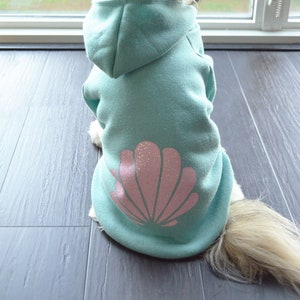 May include: A small dog wearing a light blue hoodie with a pink seashell design on the back. The dog is facing away from the camera, sitting on a dark gray floor near a window.