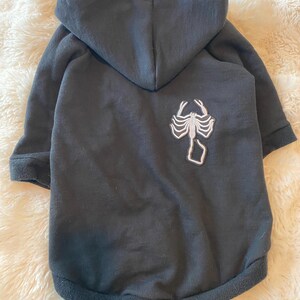May include: A black dog hoodie with a hood and long sleeves. A white scorpion graphic is on the back. The hoodie is made of a soft material.