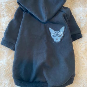 May include: Black dog hoodie with a hood and a patch featuring a cat's head with a pentagram symbol. The hoodie has a soft, fleece-like texture and ribbed cuffs and hem. The patch is light blue and gray.