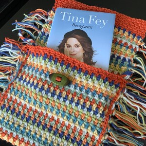 May include: A crocheted bag with colorful stripes and fringe, featuring a button closure. The bag is holding a book titled "Bossypants" by Tina Fey.