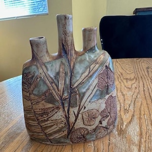 May include: A handcrafted ceramic vase with three spouts. The vase features a textured surface with a design of leaves and botanical elements in shades of brown, green, and beige. The vase is a unique decorative piece.