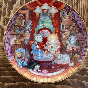 May include: A decorative ceramic plate with a red background and gold trim. The plate features a whimsical illustration of cats having tea in a cozy room. The cats are dressed in clothing and holding teacups. The plate has the word "Love" written on a heart.