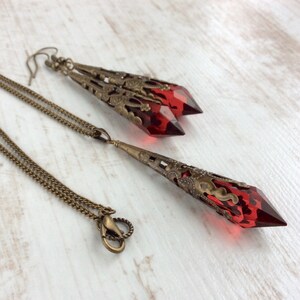 May include: A jewelry set featuring a necklace and earrings. The set has a bronze-colored chain and filigree metalwork. The earrings and pendant feature faceted, red-colored crystal-like stones. The jewelry is displayed on a light-colored wooden surface.