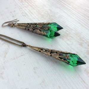 May include: A jewelry set featuring a necklace and earrings. The necklace has a long, bronze-colored chain with a pendant of a faceted, emerald-green crystal. The earrings have a similar design, with green crystals and bronze filigree.