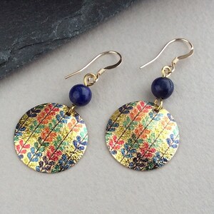 May include: Gold-tone earrings with a circular design featuring colorful leaf patterns. Each earring has a small, dark blue bead and a gold-colored hook. The earrings are displayed on a light gray surface.