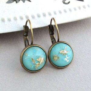 May include: A pair of round turquoise earrings with gold leaf accents. The earrings have a bronze-colored metal frame and a lever-back closure. The earrings are displayed on a white decorative surface.