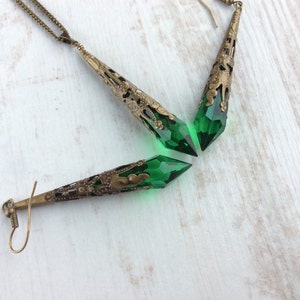 May include: A vintage-style jewelry set featuring a necklace and earrings. The set includes a long, ornate pendant and two earrings with a bronze-colored metal filigree design. Each piece holds a faceted, emerald-green crystal, creating a striking contrast.