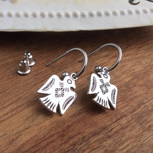 May include: Silver-coloured earrings with a bird motif, displayed on a wooden board. The earrings have a detailed etched design. The background includes a decorative metal piece. The earrings are suspended from a hoop.