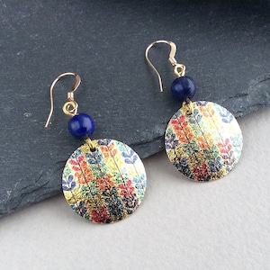 May include: A pair of gold-toned dangle earrings. Each earring features a round, patterned pendant with gold, red, blue, and yellow hues, paired with a small, dark blue bead. The earrings hang from gold-colored hooks.