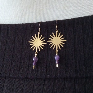 May include: Gold-colored sunburst earrings with a dangling cluster of purple beads. The earrings are suspended from a simple hook and are displayed on a white surface against a black ribbed fabric.