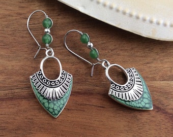 Puede incluir: Pendientes plateados con un colgante verde texturizado en forma de punta de flecha. Los pendientes tienen un borde plateado decorativo y están adornados con cuentas verdes. Los pendientes se exhiben sobre una superficie de madera.