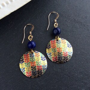 May include: Gold-tone earrings with a circular design featuring a colorful leaf pattern. Each earring has a dark blue bead and a gold hook. The earrings are displayed on a dark surface.