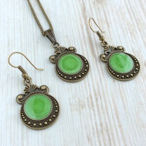 May include: A jewelry set featuring a necklace and earrings. The set has a bronze-colored metal with a decorative design. Each piece has a round, bright green center. The earrings have a hook closure.