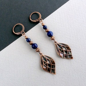 May include: A pair of copper-coloured drop earrings. Each earring features a leaf-shaped charm with a geometric pattern, accented by dark blue beads and copper-coloured metal beads. The earrings have lever-back closures.