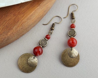 Puede incluir: Pendientes colgantes con cuentas rojas, detalles en espiral color bronce y colgantes circulares. Los pendientes se muestran sobre una superficie de madera con un elemento decorativo blanco en el fondo. Estética vintage.