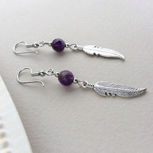 May include: A pair of dangle earrings featuring a faceted purple bead and a silver-colored feather charm. The earrings have a hook closure and are displayed on a light background. The jewelry is designed for a bohemian or nature-inspired aesthetic.