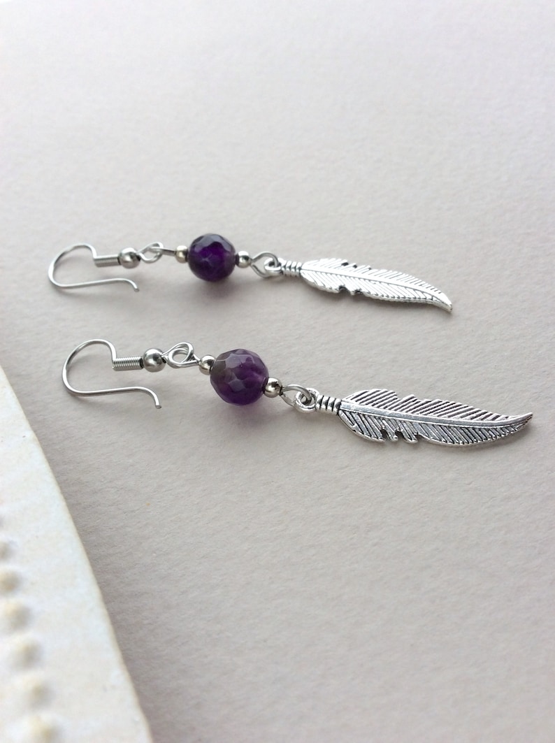 May include: A pair of dangle earrings featuring a faceted purple bead and a silver-colored feather charm. The earrings have a hook closure and are displayed on a light background. The jewelry is designed for a bohemian or nature-inspired aesthetic.