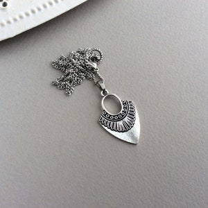 May include: Silver-toned pendant necklace with a unique design. The pendant features an oval top with circular accents and a stylized, textured bottom. The necklace chain is also silver-toned, and the background is a neutral gray.