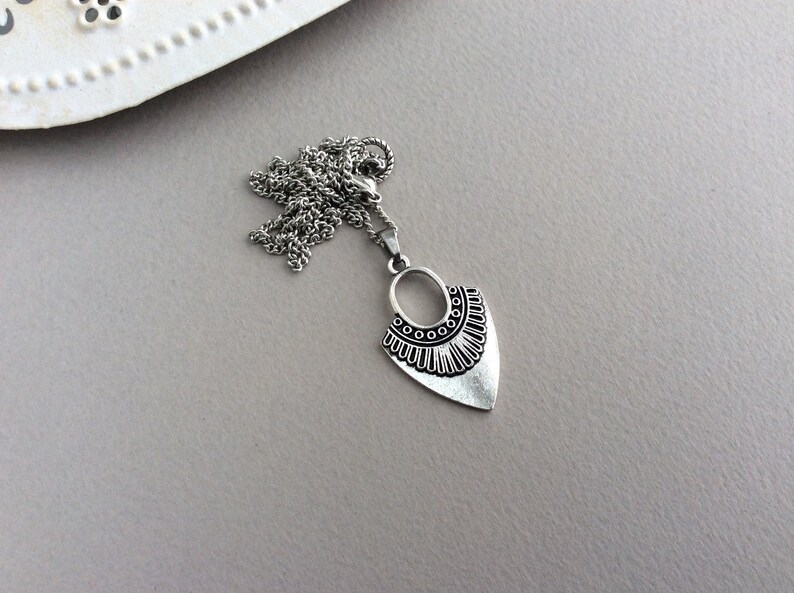 May include: Silver-toned pendant necklace with a unique design. The pendant features an oval top with circular accents and a stylized, textured bottom. The necklace chain is also silver-toned, and the background is a neutral gray.