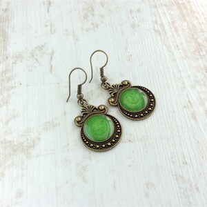 May include: A pair of antique brass-toned earrings. Each earring features a round, green, glossy center surrounded by a beaded border. The earrings have decorative filigree accents and a hook closure. The earrings are displayed on a white wooden surface.