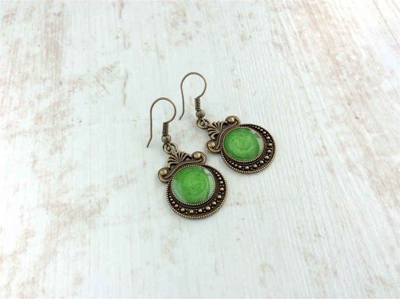 May include: A pair of antique brass-toned earrings. Each earring features a round, green, glossy center surrounded by a beaded border. The earrings have decorative filigree accents and a hook closure. The earrings are displayed on a white wooden surface.