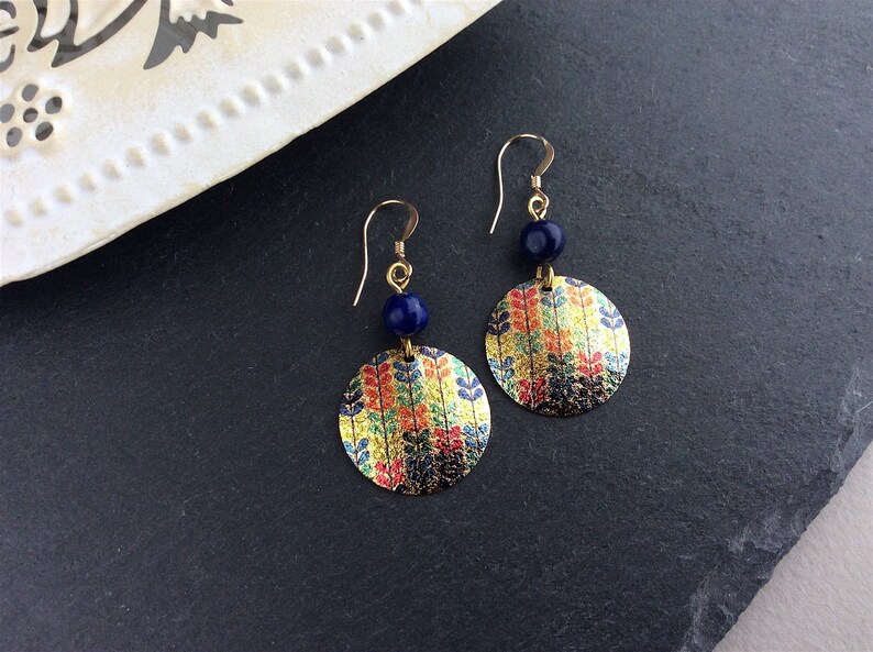 May include: Gold-tone earrings with a circular design featuring a colorful leaf pattern. Each earring has a dark blue bead and a gold hook. The earrings are displayed on a dark surface.