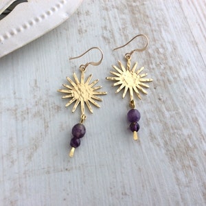 May include: Gold-tone sunburst earrings with a hammered texture. Each earring features a sun-shaped pendant with a dangling strand of two purple amethyst beads. The earrings are displayed on a light wood surface.