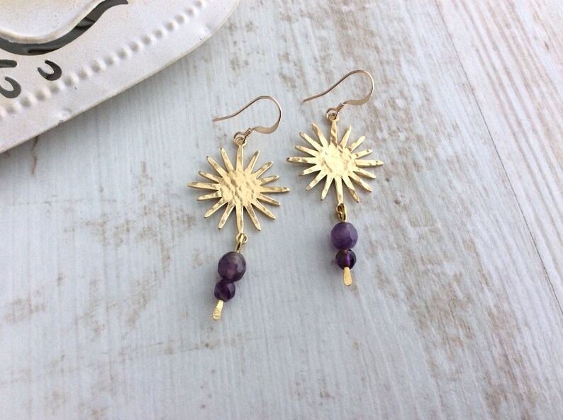 May include: Gold-tone sunburst earrings with a hammered texture. Each earring features a sun-shaped pendant with a dangling strand of two purple amethyst beads. The earrings are displayed on a light wood surface.
