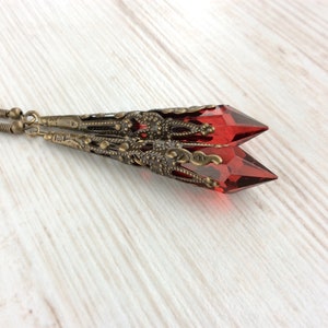 May include: A pair of long, dangle earrings featuring faceted, red crystal pendants. The crystals are encased in ornate, antique bronze filigree settings. The earrings have a vintage, gothic aesthetic.