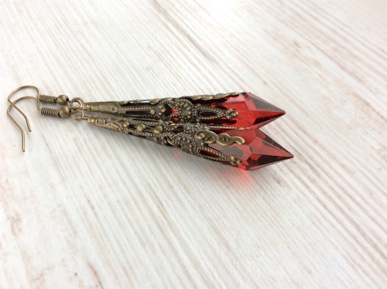 May include: A pair of long, dangle earrings featuring faceted, red crystal pendants. The crystals are encased in ornate, antique bronze filigree settings. The earrings have a vintage, gothic aesthetic.