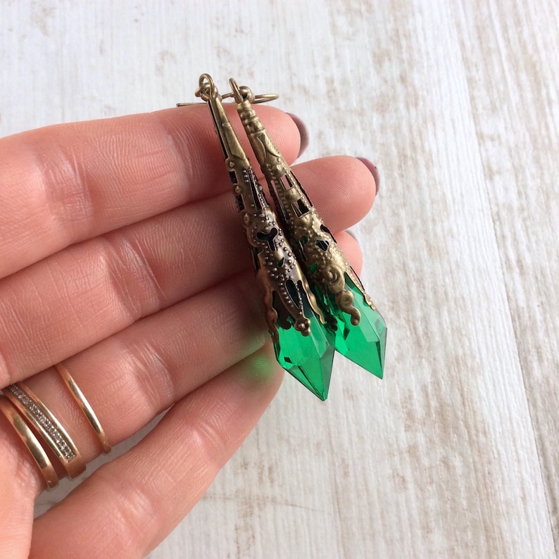 May include: A pair of long, ornate earrings with a vintage aesthetic. The earrings feature a filigree design in a bronze tone, with a vibrant green, faceted crystal at the bottom. The earrings are held in a hand.