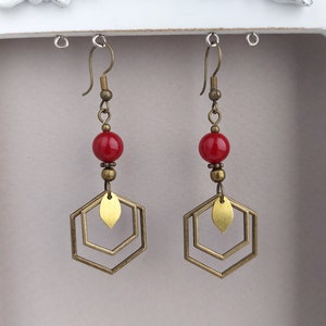 May include: A pair of dangle earrings featuring red beads, brass-colored metal accents, and geometric hexagon shapes. The earrings have a vintage aesthetic and are suspended from brass-colored ear wires.
