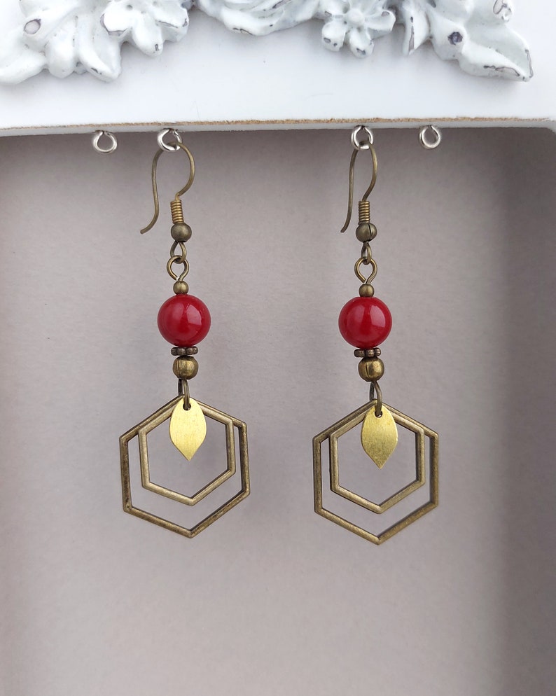 May include: A pair of dangle earrings featuring red beads, brass-colored metal accents, and geometric hexagon shapes. The earrings have a vintage aesthetic and are suspended from brass-colored ear wires.