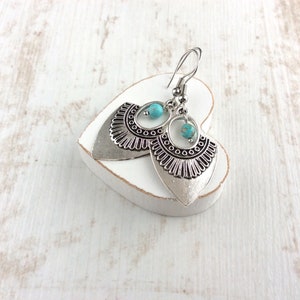 May include: Silver-toned dangle earrings with a turquoise bead accent. The earrings have a detailed, fan-shaped design with a textured finish. They are displayed on a white heart-shaped object against a white wooden background.