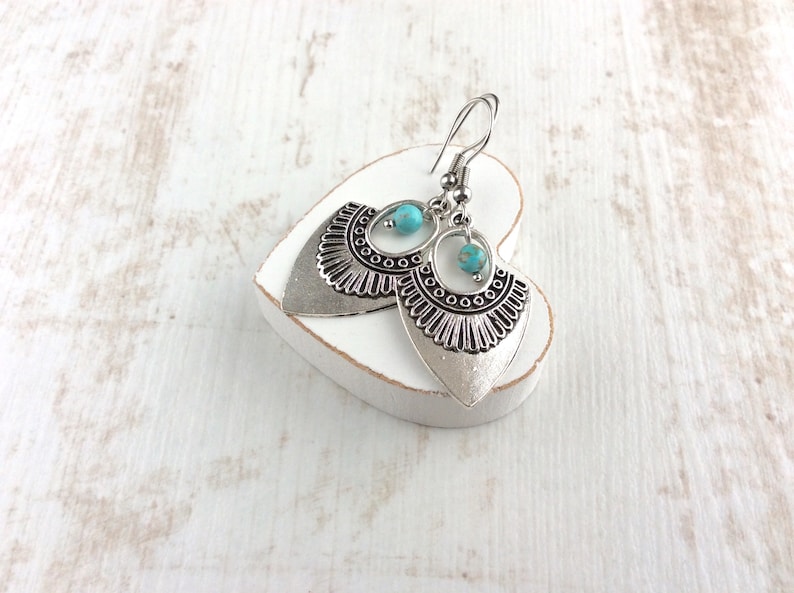 May include: Silver-toned dangle earrings with a turquoise bead accent. The earrings have a detailed, fan-shaped design with a textured finish. They are displayed on a white heart-shaped object against a white wooden background.
