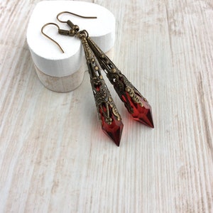 May include: A pair of long, ornate earrings with a vintage aesthetic. The earrings feature a deep red, faceted crystal pendant encased in a filigree metal setting. The metal has a warm, antique brass finish, and the earrings hang from simple ear wires.