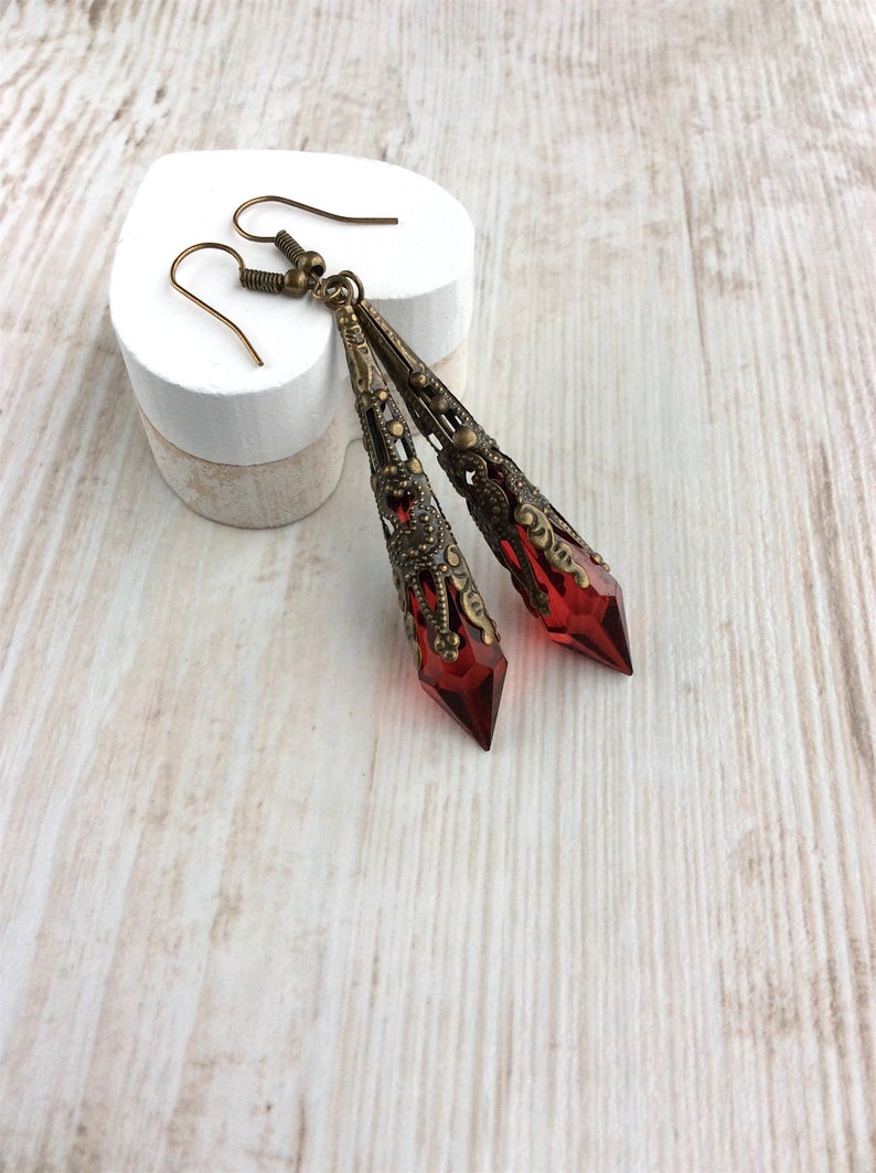May include: A pair of long, ornate earrings with a vintage aesthetic. The earrings feature a deep red, faceted crystal pendant encased in a filigree metal setting. The metal has a warm, antique brass finish, and the earrings hang from simple ear wires.