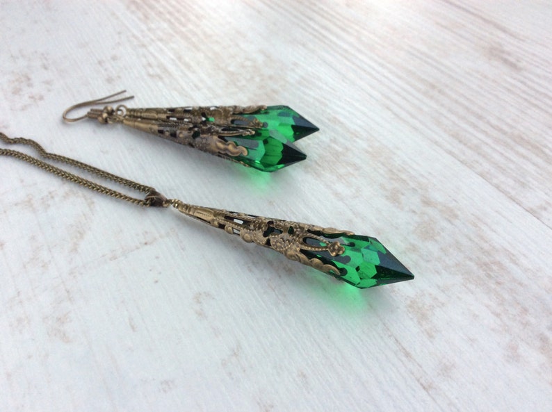 May include: A jewelry set featuring a necklace and earrings. The necklace has a long, bronze-colored chain with a pendant of a faceted, emerald-green crystal. The earrings have a similar design, with green crystals and bronze filigree.