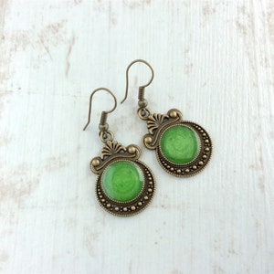 May include: A pair of antique-style earrings with a green center. The earrings have a bronze-colored metal frame with decorative filigree and a beaded border. The green center has a glossy finish.