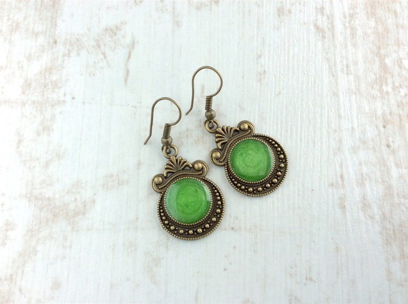 May include: A pair of antique-style earrings with a green center. The earrings have a bronze-colored metal frame with decorative filigree and a beaded border. The green center has a glossy finish.