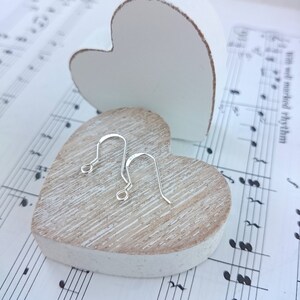 May include: A pair of silver-colored, curved fish hook earrings rest on a light-colored, heart-shaped wooden block. A white heart-shaped object is in the background, and the background is sheet music.