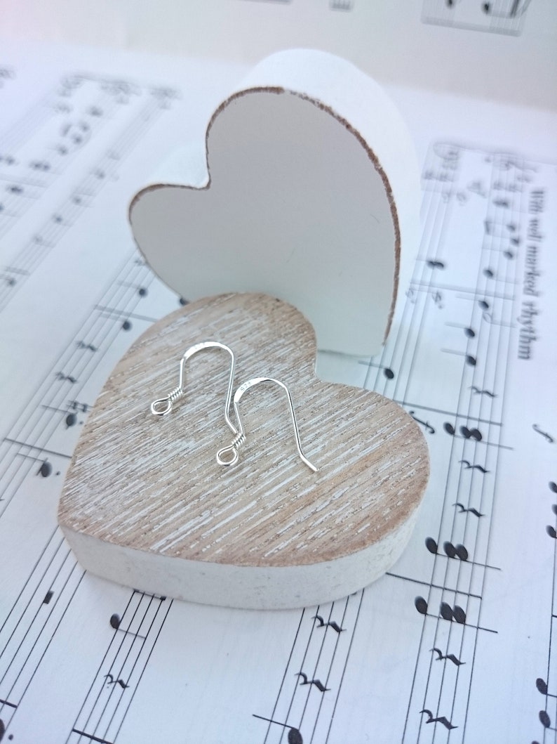 May include: A pair of silver-colored, curved fish hook earrings rest on a light-colored, heart-shaped wooden block. A white heart-shaped object is in the background, and the background is sheet music.
