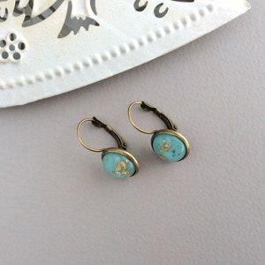 May include: A pair of leverback earrings with oval turquoise-colored stones. The stones are flecked with gold leaf and set in antique bronze-toned metal. The earrings are displayed on a light gray surface.