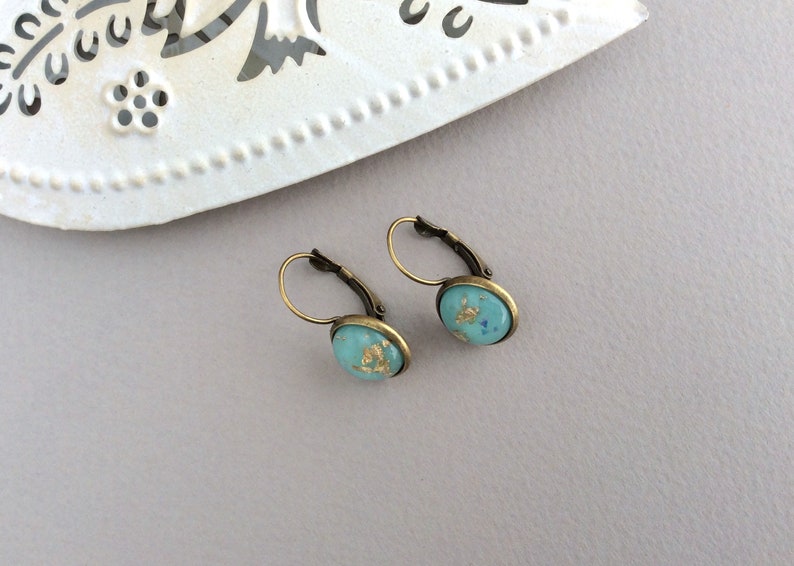 May include: A pair of leverback earrings with oval turquoise-colored stones. The stones are flecked with gold leaf and set in antique bronze-toned metal. The earrings are displayed on a light gray surface.