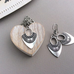 May include: A silver-toned jewelry set featuring a necklace and earrings. The necklace pendant and earrings have a shield-like shape with an oval top and a decorative, patterned bottom. The set is displayed on a wooden heart.