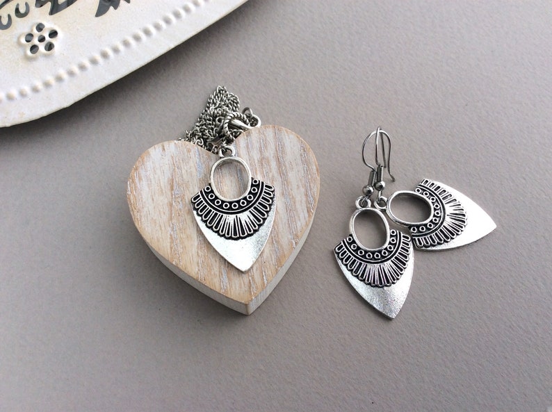 May include: A silver-toned jewelry set featuring a necklace and earrings. The necklace pendant and earrings have a shield-like shape with an oval top and a decorative, patterned bottom. The set is displayed on a wooden heart.